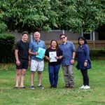 A group of five people standing outdoors under a large tree, smiling at the camera. Two individuals in the center are holding partnership documents.