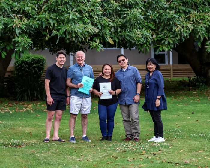 A group of five people standing outdoors under a large tree, smiling at the camera. Two individuals in the center are holding partnership documents.