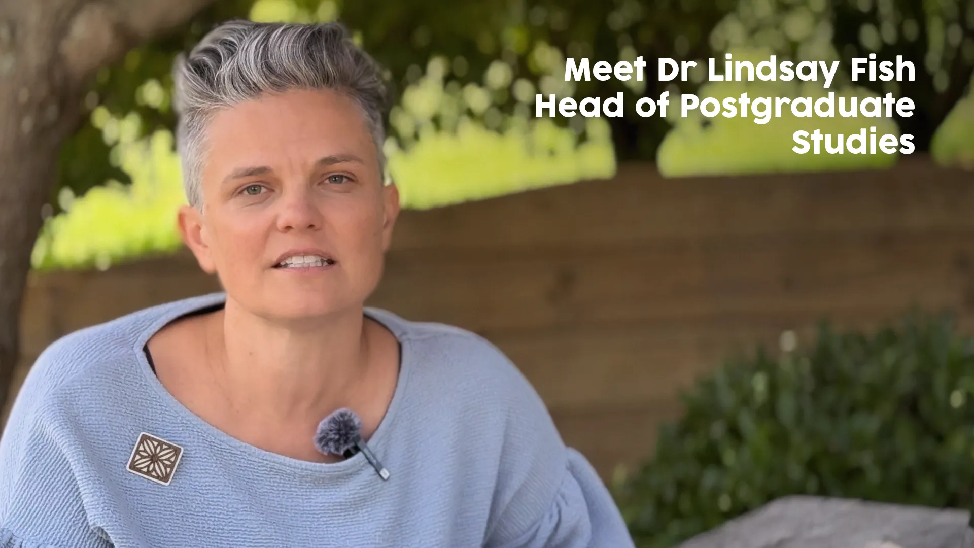 Dr Lindsay Fish, wearing a light blue sweater and a microphone, sitting outdoors with greenery and a wooden fence in the background, introducing herself as Head of Postgraduate Studies.