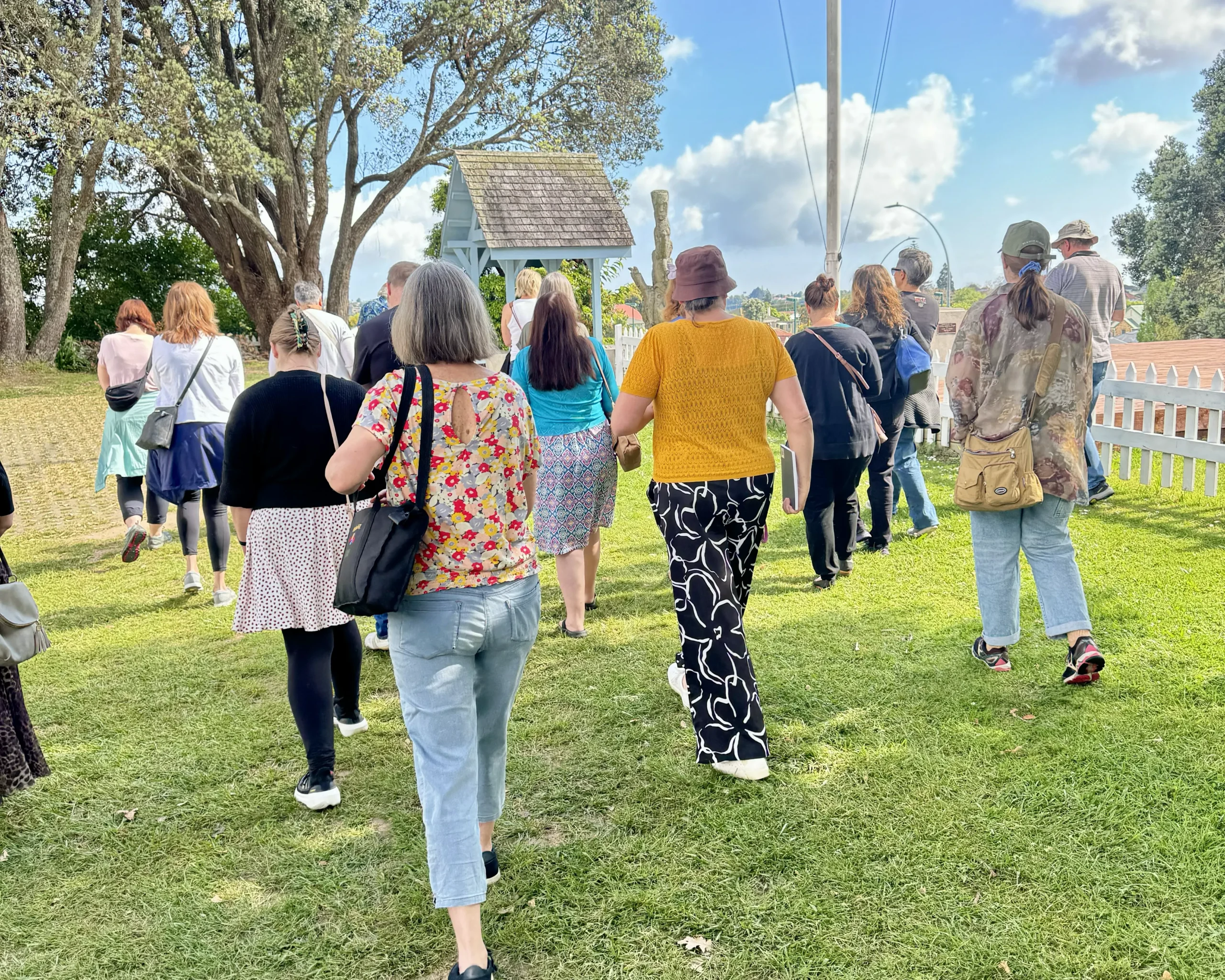 A group of people walking through a grassy area under large trees at the Pukehinahina battle site, with a wooden structure and a white picket fence in the background.