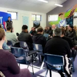 A group of people seated in a classroom setting, listening attentively to a speaker in front of a 'Teacher Education & Social Practice' banner. The room is decorated with a large Māori-inspired mural featuring a cross.