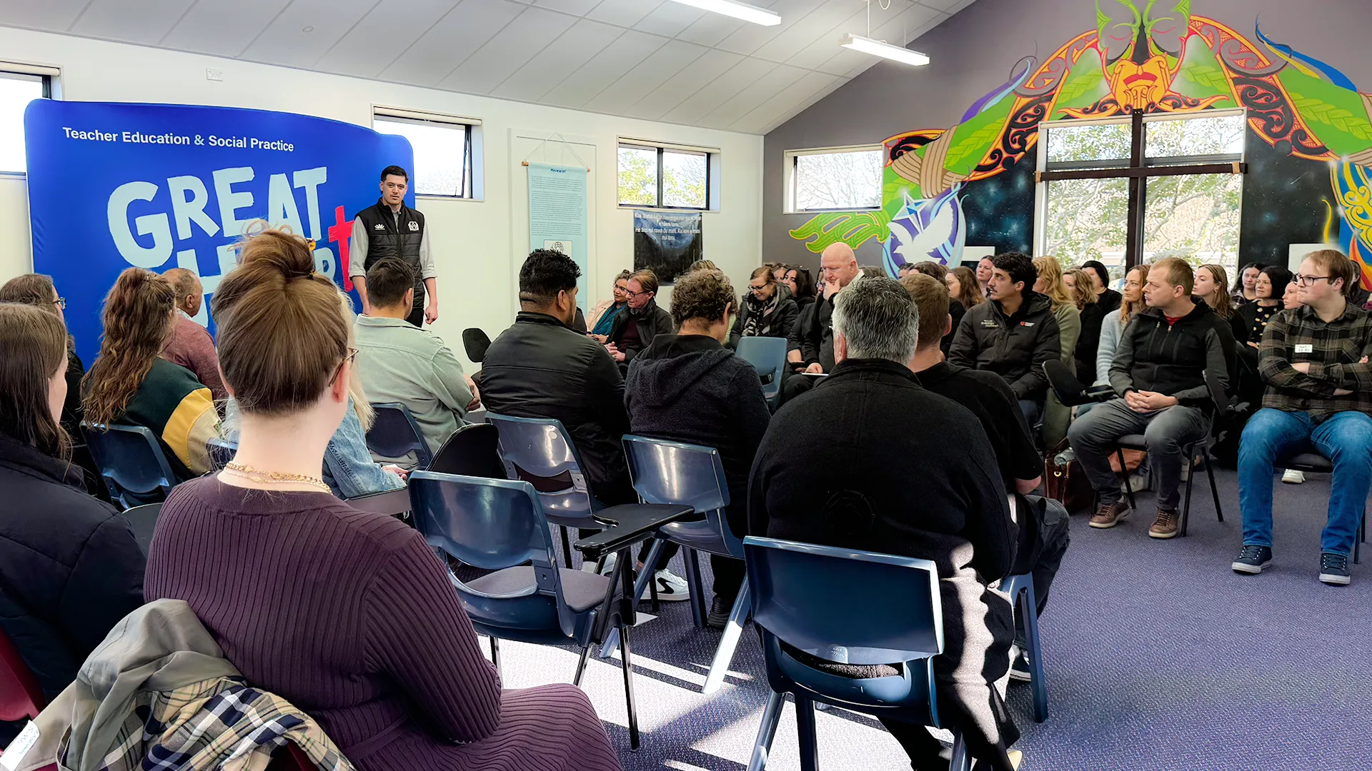 A group of people seated in a classroom setting, listening attentively to a speaker in front of a 'Teacher Education & Social Practice' banner. The room is decorated with a large Māori-inspired mural featuring a cross.